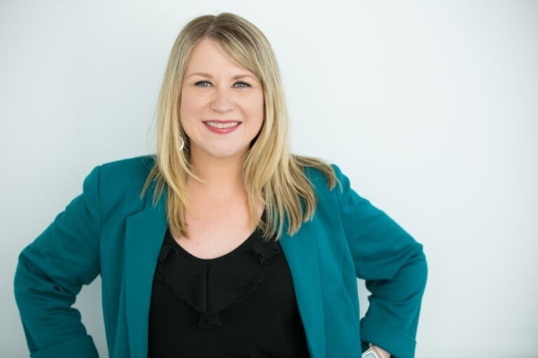 A photo of a woman with blonde hair wearing a black shirt and teal blazer.