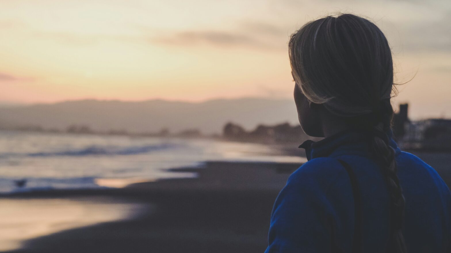 A photo of a woman standing on a beach looking at the ocean at sunset. The lighting is very dim.