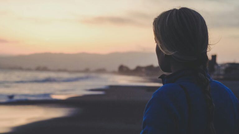 A photo of a woman standing on a beach looking at the ocean at sunset. The lighting is very dim.